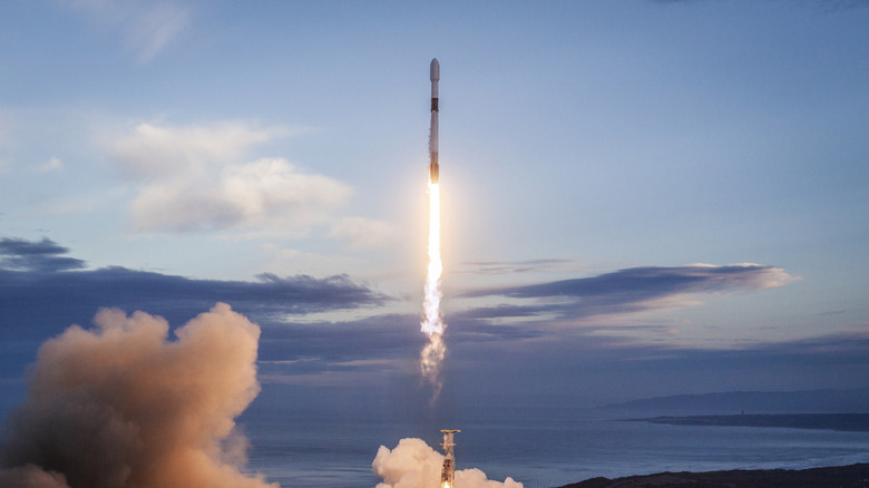 a spacecraft takes off, jets of flame visible on a backdrop of clouds and blue sky