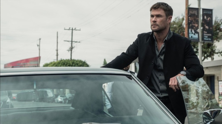 Jewel thief Mike Davis (Chris Hemsworth), clothed in a slick black suit, stands outside the propped door of his getaway car in Crime 101