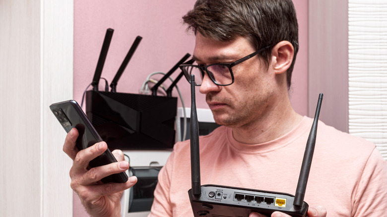 Person holding a phone in one hand and a router in the other, with a second router in the background
