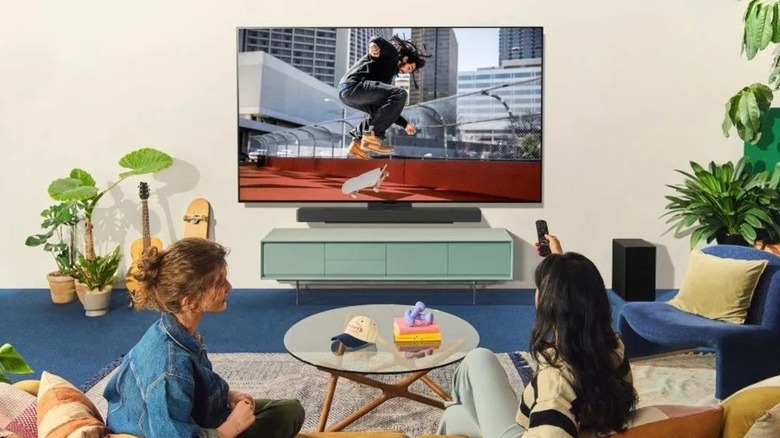 Two individuals sitting on a couch in a room, one has a remote in their right hand and is pointing it at an LG OLED C5 Smart TV