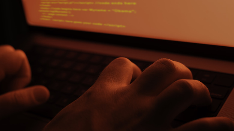 An ominous, bright red glow shines over a hackers' fingers as they type on a laptop.