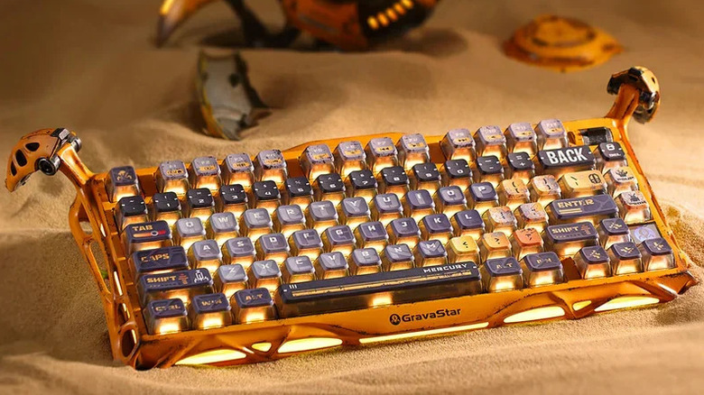 Gravastar's Mercury K1 Pro keyboard in "battle-worn yellow" staged on a bed of sand.