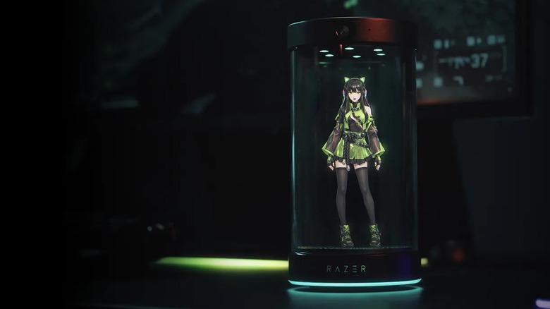 A holographic anime girl character stands in a tall glass cube with the label RAZER on the base. In the background is a dim computer monitor and green glow.