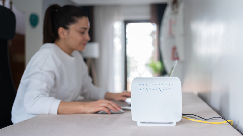 A wireless router sitting atop a desk with someone browsing a laptop in the background.