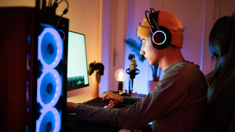 Person gaming at a desk with RGB-lit PC fans, headphones, and warm ambient lighting.