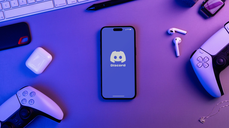 A smartphone displays the Discord logo as it lays amongst a variety of electronics, including PS5 controllers and a Apple AirPods