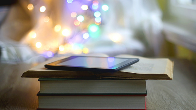 A Kindle device sitting on a stack of books with blurred string lights in the background