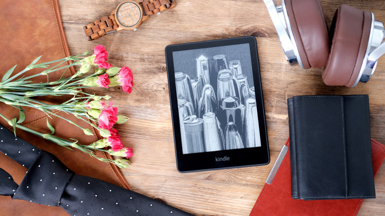 A backlit Kindle device sitting on a table with headphones, notebooks, flowers, a tie, and a watch