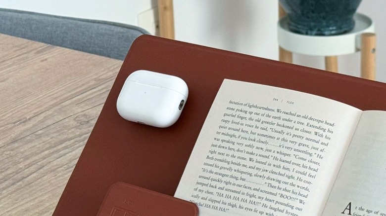 A white Apple AirPods Pro case with a book next to it, both placed on a brown stand.