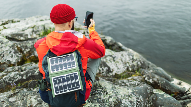 A person with a solar power bank attached to the backpack charging his phone on a cliff