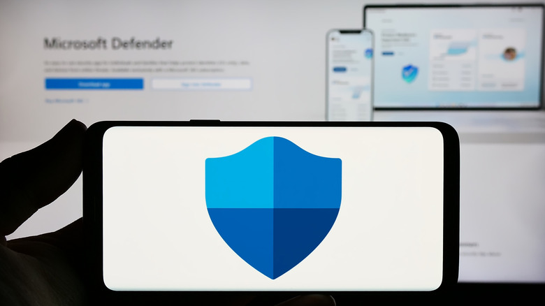 A phone displaying the Microsoft Defender logo, with the product page for Microsoft Defender Antivirus for Windows in the background