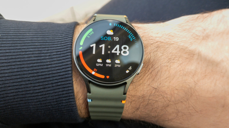 A Galaxy Watch on someone's wrist, showing time, weather, steps, heart rate, and battery life.