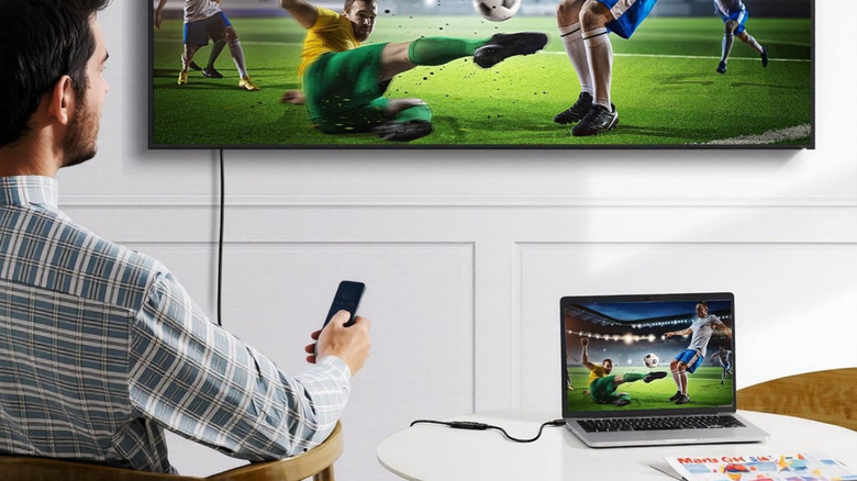A man watching a soccer game on a wall-mounted TV. A laptop is connected to the TV via an HDMI coupler.