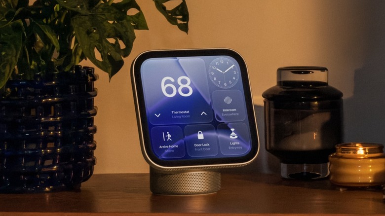 An Apple HomeView smart home hub sits on a table next to a potted plant and some candles, the HomeView is displaying its Home screen, which shows widgets for several devices as well as the time and thermostat reading