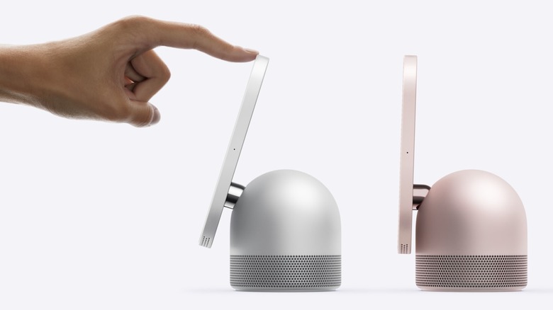 A side view of silver and rose pink Apple HomeView smart home hubs, a user's index finger on their left hand is touching the top of the screen on the silver HomeView