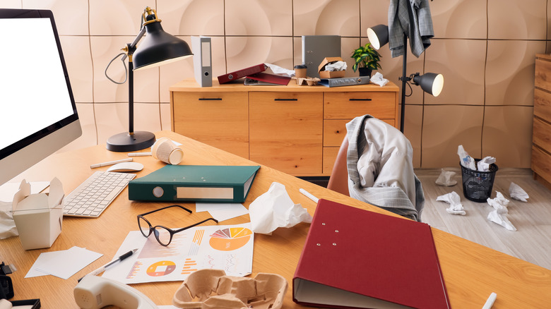 A messy office space littered with papers, binders, takeout containers, clothes, and garbage