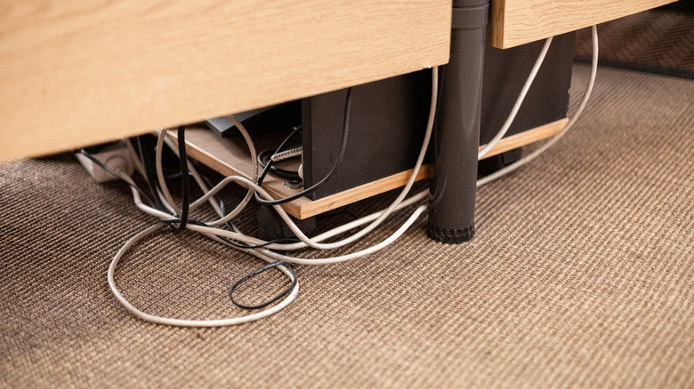 Disorganized cables showing underneath a desk on a beige carpet