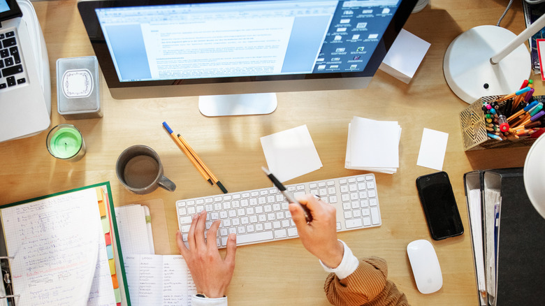 Two hands hovering over a desk cluttered with papers, pens, and various electronics in front of a computer monitor