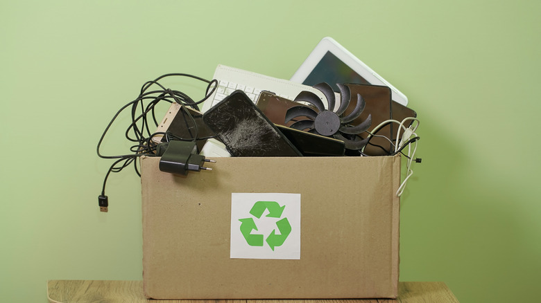 Worn-looking gadgets and tech accessories in a brown box with a green recycling symbol on it against a solid green background