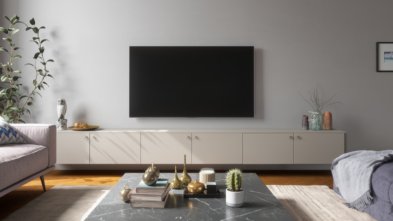 A television with a black screen hangs from a wall in a contemporary room containing a long white TV stand, a couch, and a table that has assorted brick-a-brack