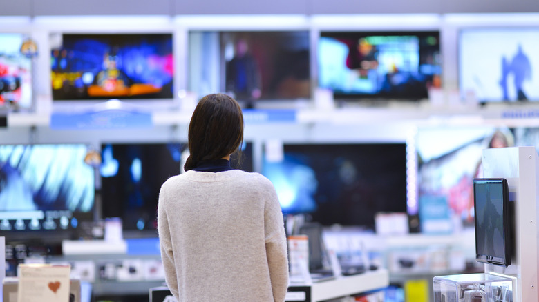 An individual with black hair and a gray shirt stands before a wall of televisions in what appears to be a shopping center