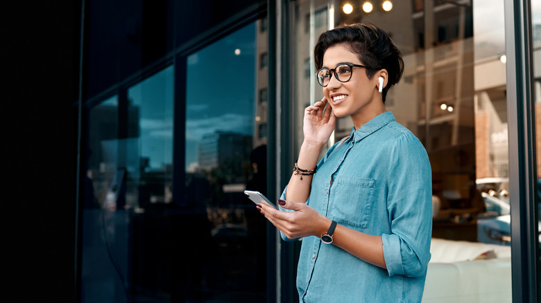 A woman standing outside a building holding a smartphone and listening to something through earbuds