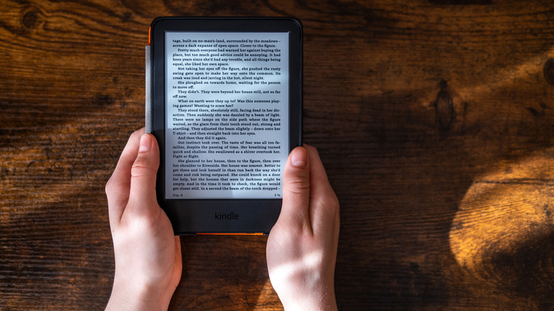 Two hands holding a Kindle Paperwhite in a case, with the screen on, against a wooden background