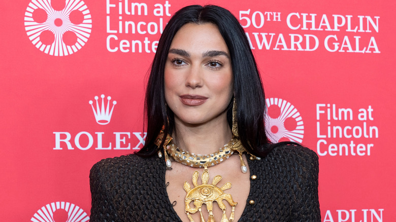 Singer-songwriter Dua Lipa attending an Award Gala, she is dressed in black and is wearing a large gold necklace that appears to have a eyeball-shaped design