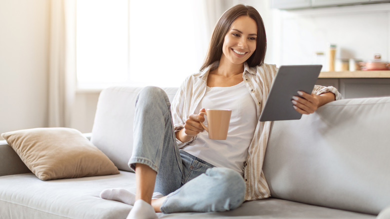 A dark haired woman in a white shirt and jeans drinks coffee while reading from a tablet on her couch.