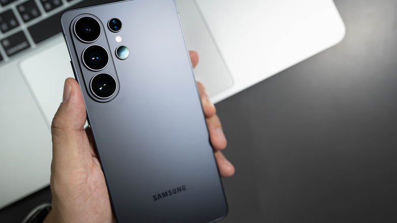 A hand holding a Samsung Galaxy S26 smartphone above a laptop.