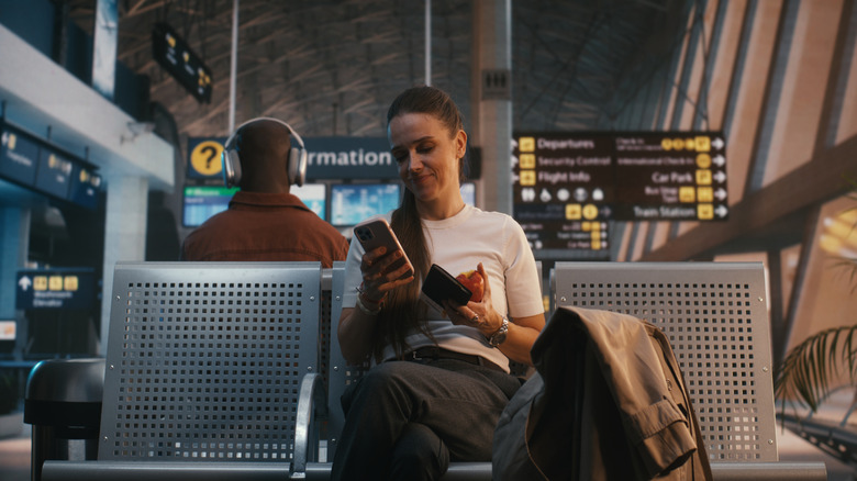 A woman smiles at her iPhone while sitting in an airport terminal.