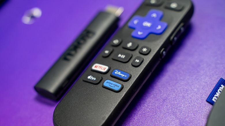 Close up of a Roku streaming stick and a Roku brand remote on a purple background.