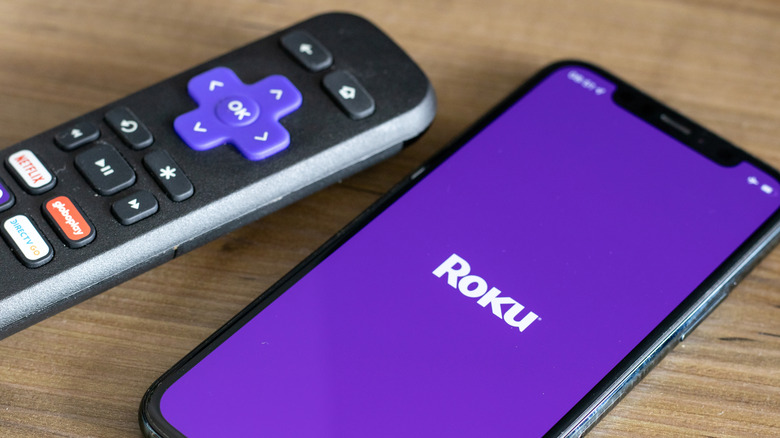 Roku brand remote sitting next to a smartphone with the Roku logo onscreen.