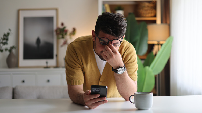 A man in a yellow shirt worryingly covers his eyes while staring at his phone screen.