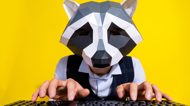 A man with a racoon mask types furiously on a keyboard before a bright yellow background