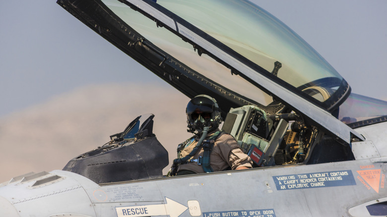 A pilot in the cockpit of an F-16 fighter jet with the canopy open.