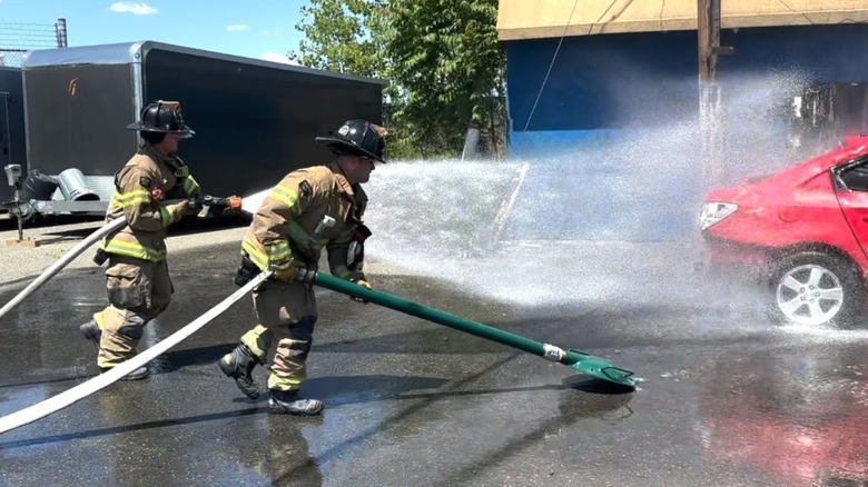 Two firefighters showcasing how to treat car fires. One is spraying water with a hose with the other holds a green pole connected to a turtle-shell looking object on one side and the water supply on the other