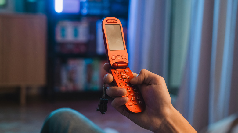 A hand holding a red-orange flip phone with a blurry living room in the background.