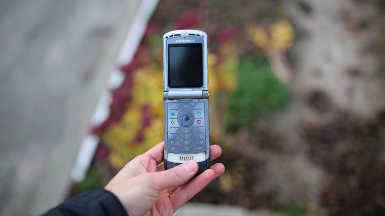 A silver Motorola Razr V3 held in someone's hand with a blurred background of a curbside.