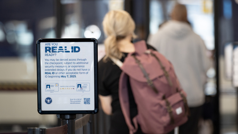 A sign at the airport asking travelers if they are Real ID compliant, with as explanation as to what may happen if someone is lacking proper identification, an individual with a mauve backpack is facing away from the sign in the background