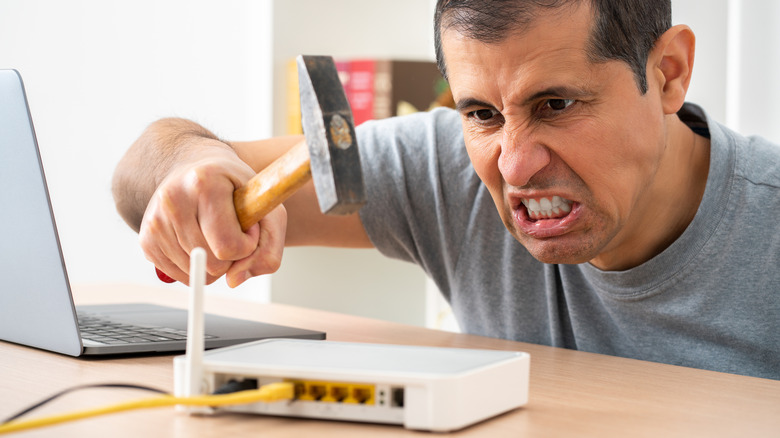 An angry man takes a hammer to his internet Wi-Fi router besides his open laptop.