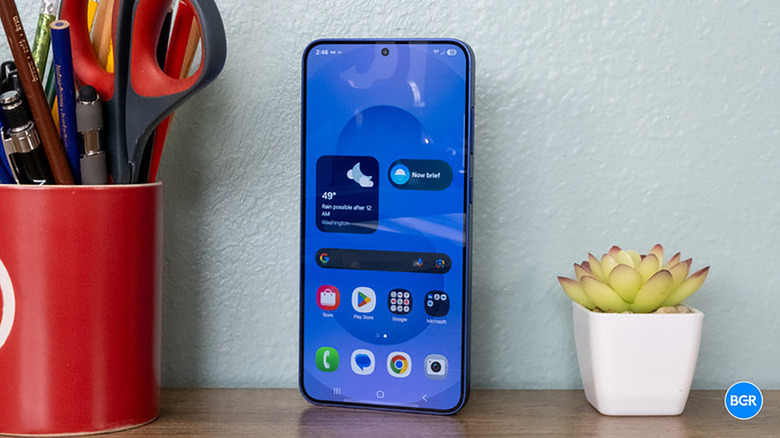 The Samsung Galaxy S25 propped up on a desk beside a plant and a cup with pens.