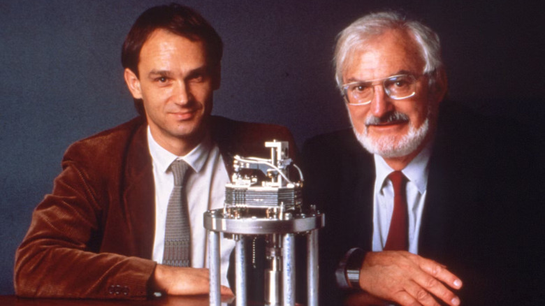 Gerd Binnig and Heinrich Rohrer sit behind the scanning tunneling microscope they invented, resting on a table.