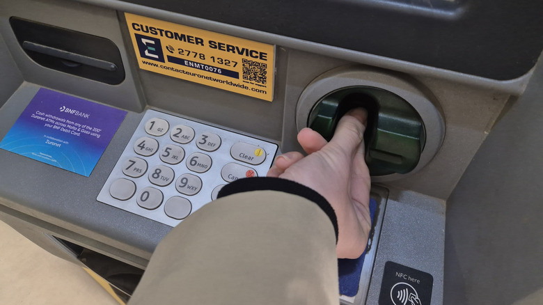 A person inserts their hand into an ATM card slot while standing at a machine, with the keypad, contactless payment symbol, and customer service information visible nearby.
