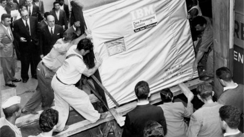 A black-and-white photo shows a group of workers using ramps to move a large, covered IBM machine into a building, while a crowd of onlookers gathers nearby on the street.