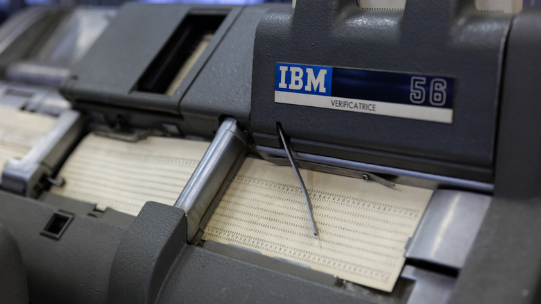 A close-up of an IBM 56 Verificatrice punch card machine feeding a punched card through its mechanism, with metal rollers and components visible.