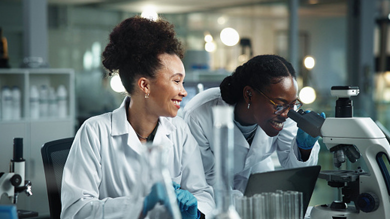 two smiling scientists work in a lab, looking through a microscope