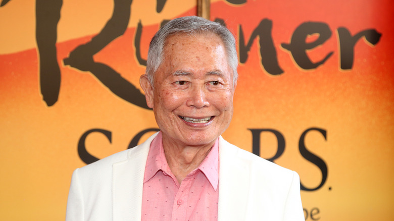 George Takei smiling in front of orange sign