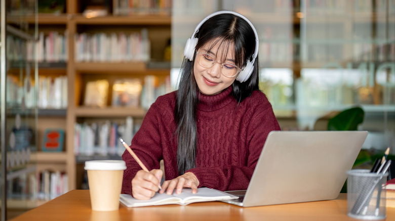 A woman sitting at a table in a library writing in a journal with a laptop and coffee cup in front of her. She is wearing a red sweater, glasses, and white headphones.
