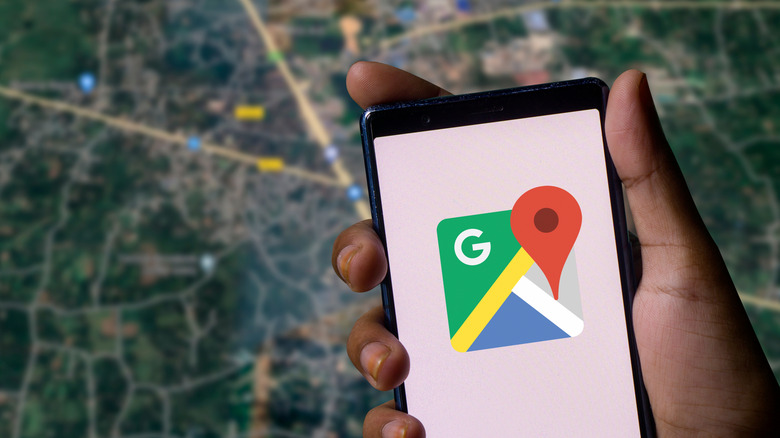 A closeup of an individual's right hand holding a smartphone displaying the Google Maps logo, behind the phone is a slightly blurry map acting as a backdrop